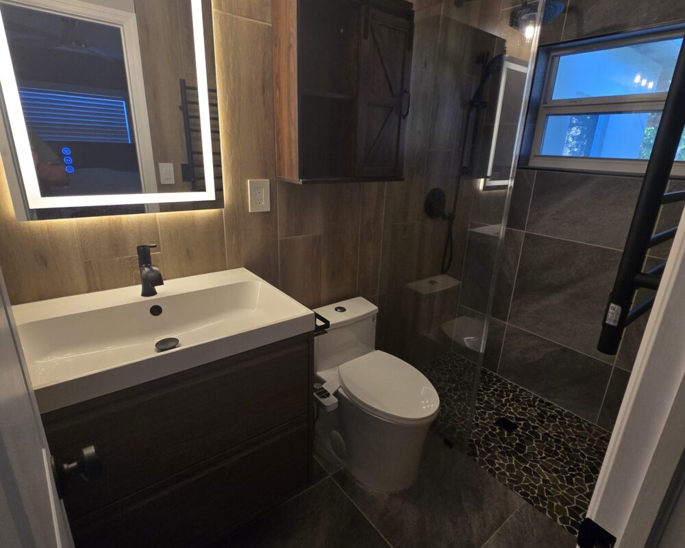 Modern bathroom remodel featuring a sleek vanity with a white sink, black faucet, and illuminated mirror, alongside a stylish shower with glass enclosure and pebble flooring, showcasing TurnKey Builds' craftsmanship.