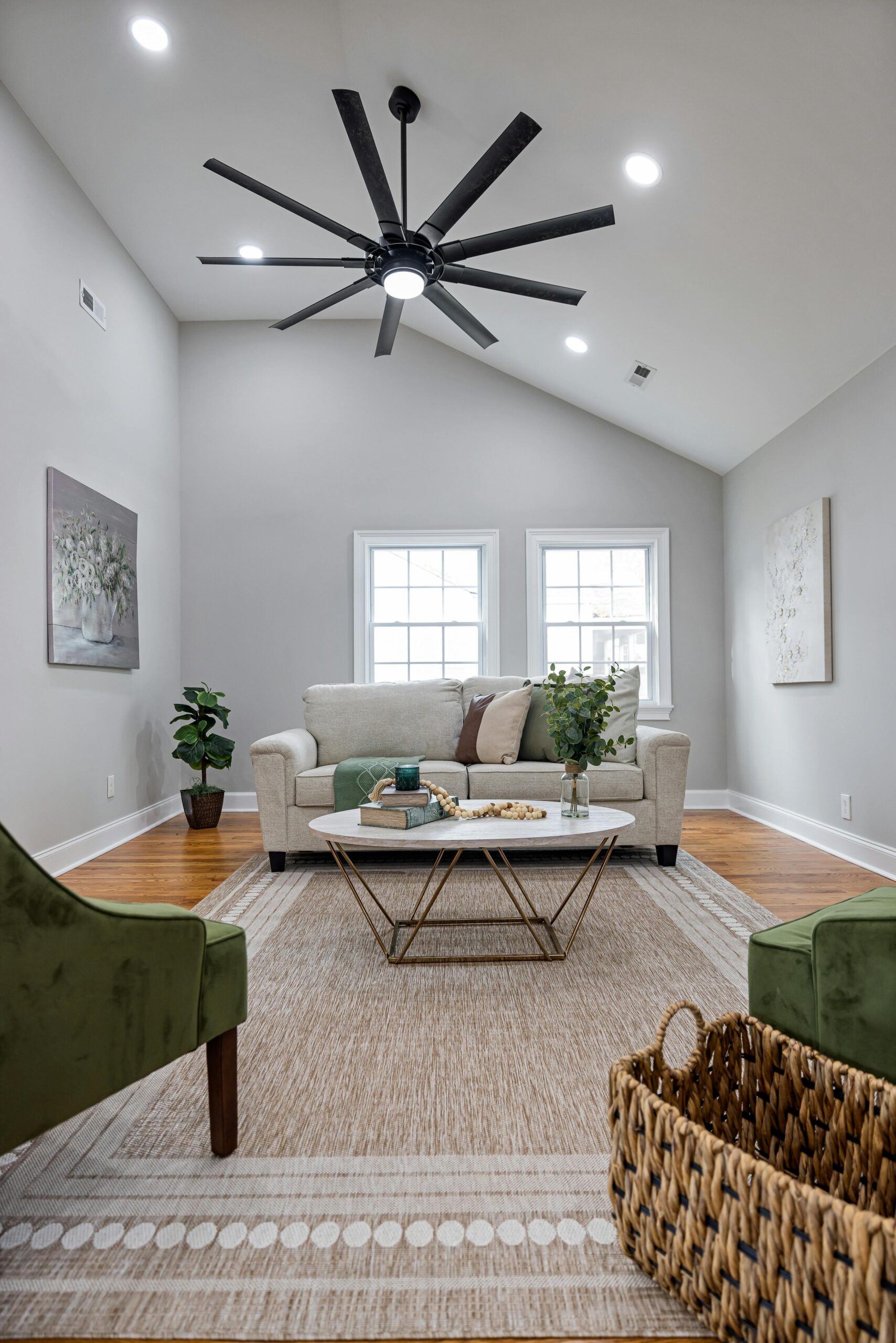Modern living room with a beige sofa, green accent chairs, a round coffee table, decorative plants, and a ceiling fan, showcasing a stylish and inviting interior design.