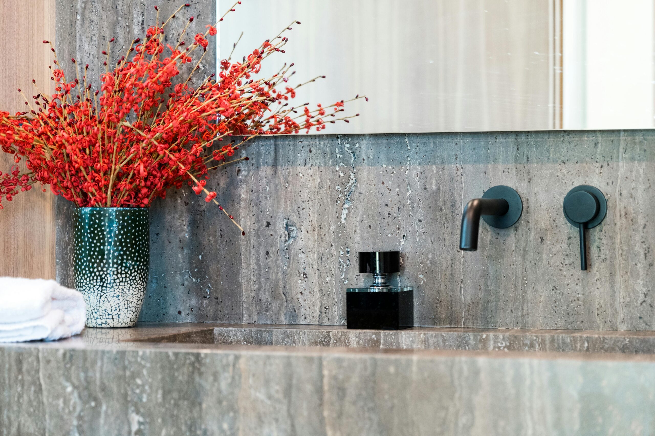 Modern bathroom vanity with black faucet fixtures, decorative gray stone surface, and a vibrant red floral arrangement in a patterned vase, showcasing luxury design elements for a powder room renovation.