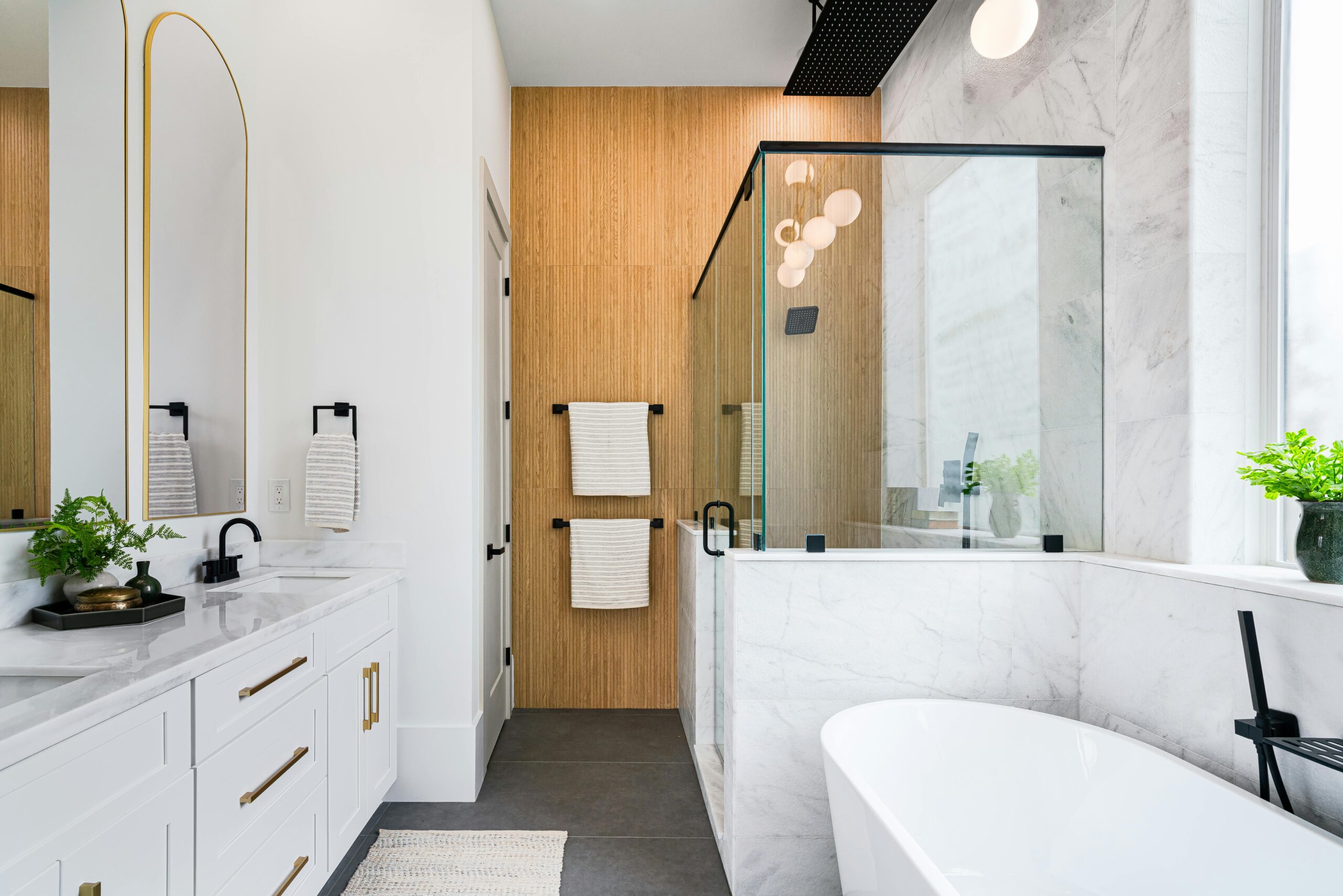Modern master bathroom featuring a freestanding tub, walk-in shower with glass enclosure, double vanity with gold accents, and natural wood wall paneling, emphasizing luxury design and functionality.