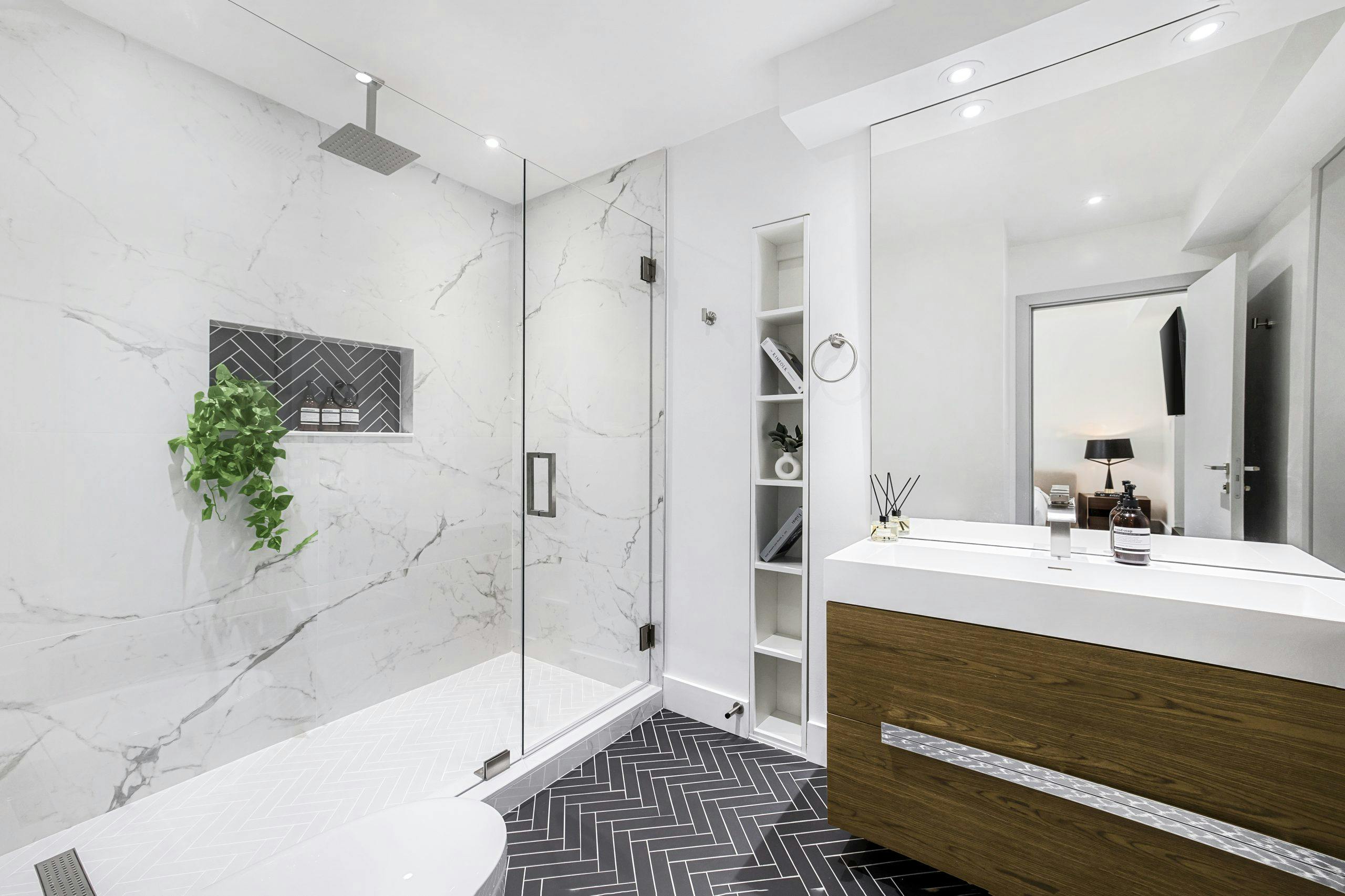 Modern bathroom design featuring a glass walk-in shower with marble walls, herringbone-patterned flooring, custom wooden vanity with sleek hardware, and stylish decor elements.