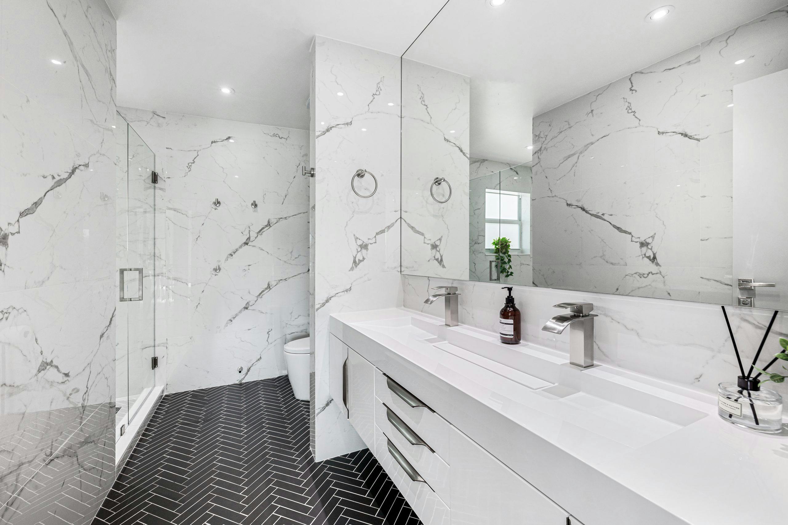 Modern bathroom with marble walls, sleek white vanity, dual faucets, and herringbone black tile flooring, showcasing luxury bathroom remodeling by TurnKey Builds.