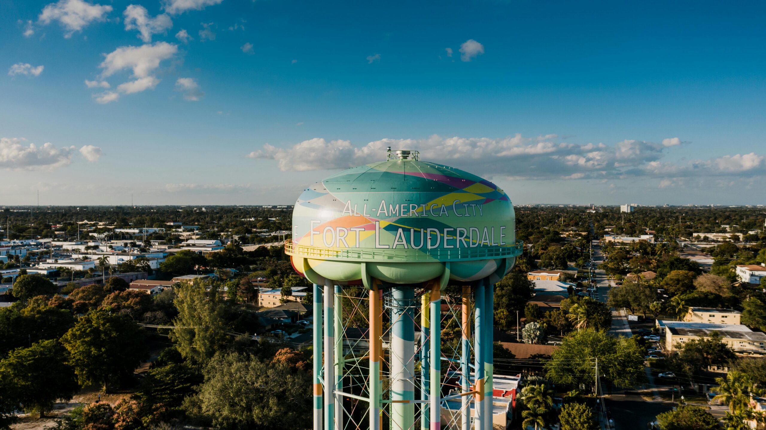 Colorful water tower with "All-America City" and "Fort Lauderdale" text, overlooking a suburban landscape in Fort Lauderdale, Florida, under a blue sky with scattered clouds.