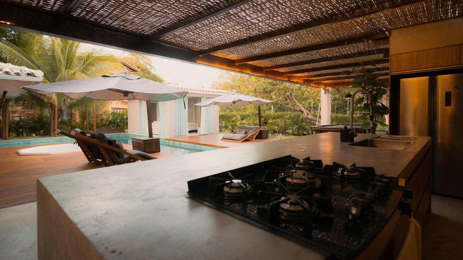 Modern outdoor kitchen with gas cooktop, overlooking a pool area featuring lounge chairs and umbrellas, surrounded by tropical landscaping, highlighting luxury renovation by TurnKey Builds in Boca Raton.
